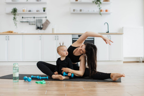 Slim brunette lady on yoga mat stretching arms and legs while looking at small kid with dumbbell in hand. Young parent and daughter in sportswear maintaining non-verbal communication during workout. - Foto, Bild
