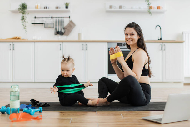 Beautiful lady and adorable child sitting with soles of feet together on floor while having fun with stretch band. Attentive baby following joyful moms instructions during physical exercises at home. - Photo, Image