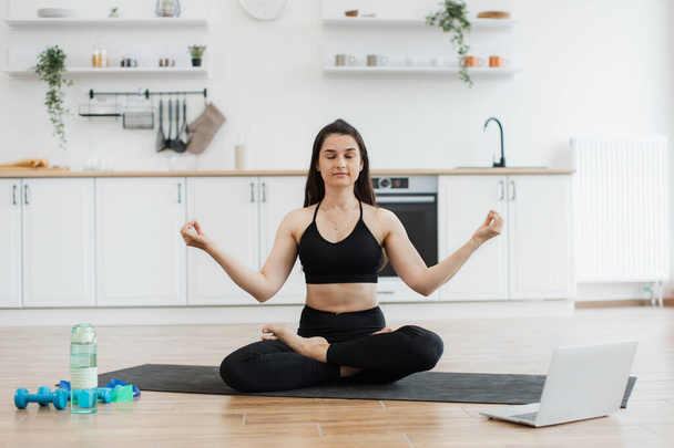Relaxed slim woman in black activewear meditating in lotus pose with hands in gyan mudra to music from laptop. Attractive lady enjoying calming tunes during yoga practice in modern apartment. - Photo, Image