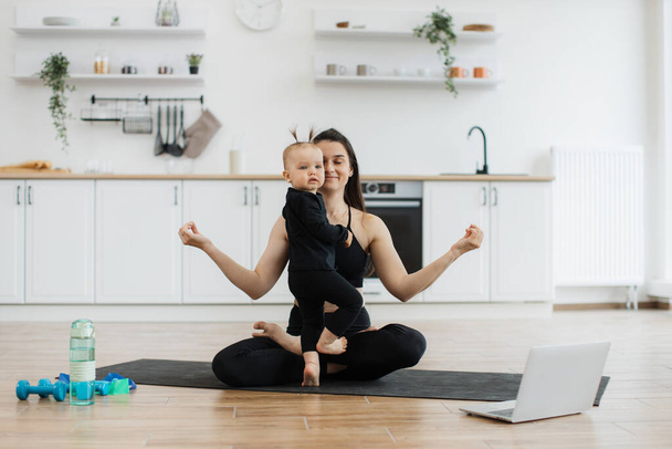 Energetic infant girl joining lady during her achieving cross-legged position due to online yoga class on device. Happy mother promoting daughters sense of calmness by meditating together at home. - Photo, image