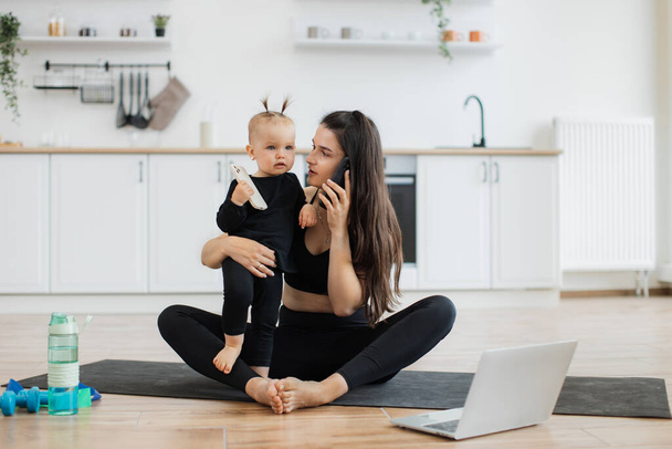 Charming woman with cell phone sitting cross-legged on yoga mat with little child on hip in kitchen interior. Busy mother speaking on mobile during exercise while cute daughter holding gadget in hand. - Photo, image