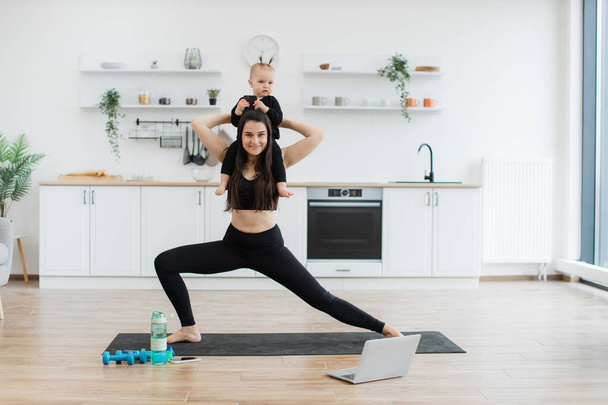 Sporty female keeping little girl on shoulders while stretching knees in standing position on mat at home. Active mommy in yoga outfit improving fitness while entertaining daughter at weekend. - Foto, afbeelding