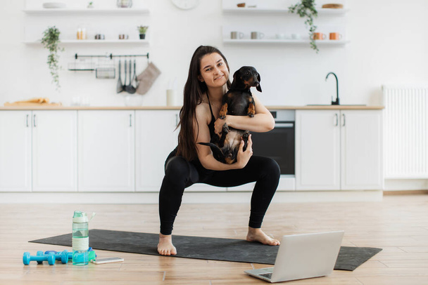 Beautiful woman in sportswear cuddling Dachshund while doing squats on rubber mat in modern apartment. Athletic person strengthening balance and coordination together with buddy pet in arms at home. - Foto, Imagem