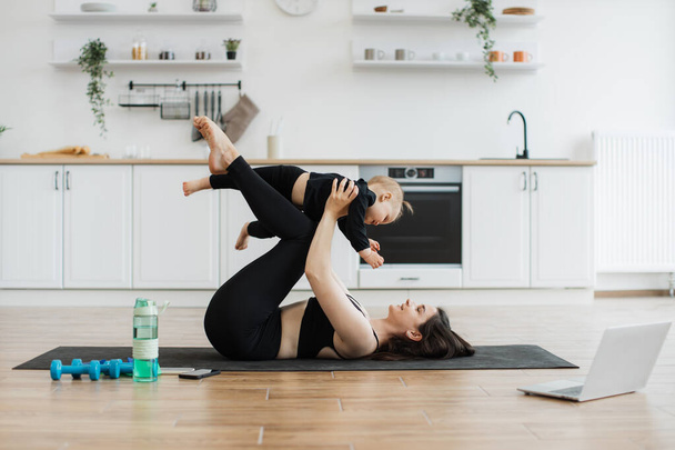 Athletic slim woman carrying infant girl on bent knees while lying on rubber mat in kitchen interior. Sporty female parent in activewear improving endurance while having fun with daughter at home. - Фото, изображение