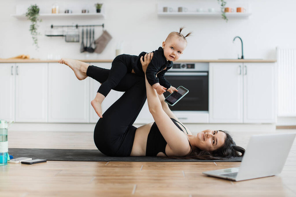 Happy young woman posing with little infant girl on shins while doing baby yoga with device in kids hand. Healthy fit mother encouraging bonding between daughter and parent without leaving home. - 写真・画像