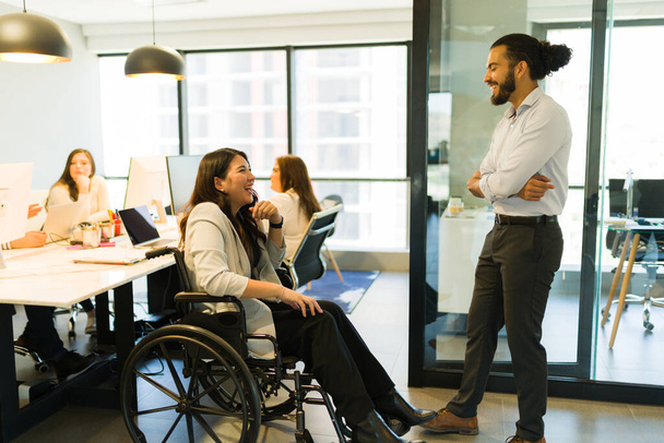 Ανάπηρος επιχειρηματίας που κάθεται σε αναπηρική καρέκλα και μιλάει με άνδρα συνάδελφο που στέκεται στο γραφείο - Φωτογραφία, εικόνα