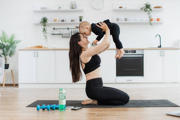 Side view of beautiful woman pressing forehead with little daughter while sitting on knees during fitness class. Endearing mother developing healthy habits at early age by involving kid to sport. - 写真・画像
