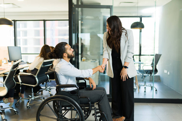 Disabled businessman sitting on wheelchair shaking hands with asian businesswoman in modern office - Photo, Image