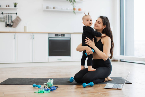 Adorable girl in black outfit standing on womans laps while trading positive energy with mom during exercises. Loving female hugging daughter with dumbbell in hand during fitness routine at home. - Foto, imagen