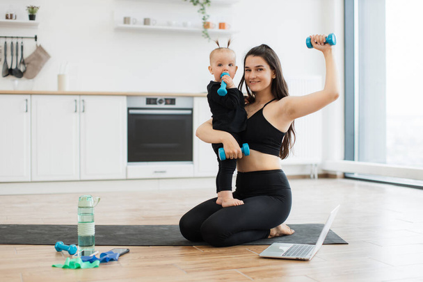 Full length portrait of sporty lady and cute little child with dumbbells sitting on yoga mat in modern kitchen. Happy mother focusing on healthy routine together with small daughter on weekend. - Fotoğraf, Görsel
