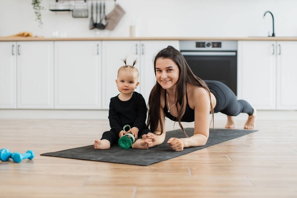 Joyful caucasian female holding plank pose on rubber mat near smiling little girl with sports bottle. Stylish mother building strong relationship with daughter while strengthening body at home. - Foto, afbeelding