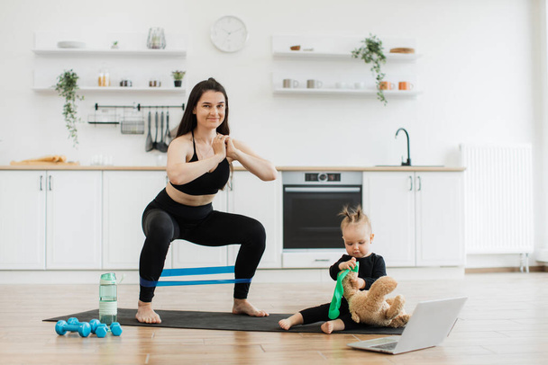 Athletic lady in sportswear practising resistance band exercises while little baby playing with teddy bear on yoga mat. Pretty brunette woman building new experience with daughter in home interior. - Zdjęcie, obraz