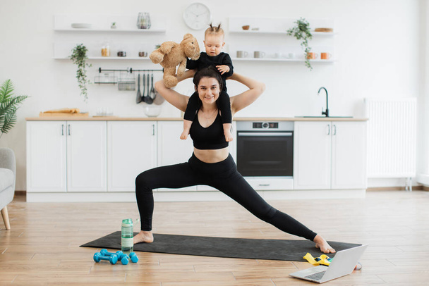 Full length portrait of joyous lady posing with infant sitting around neck with teddy bear in dining room. Healthy strong mother achieving yoga pose with double weights of daughter and soft companion. - Foto, immagini