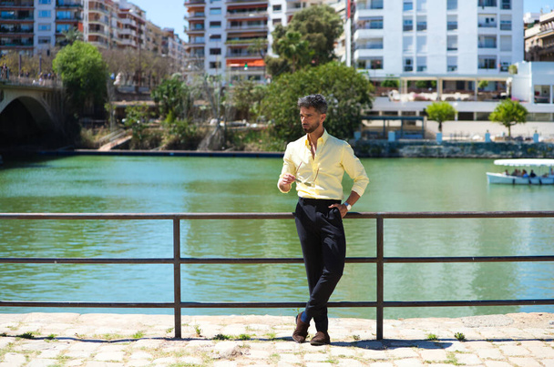 Un joven guapo con barba y gafas de sol en la mano se apoya en la barandilla del río guadalquivir en Sevilla. En la otra orilla está el famoso barrio de los artistas flamencos de Sevilla - Foto, Imagen
