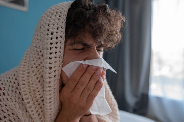 Unhappy young man in plaid suffering from fever and flu on sofa, blowing nose in napkin. Treatment of illness, cold and runny concept - Photo, Image