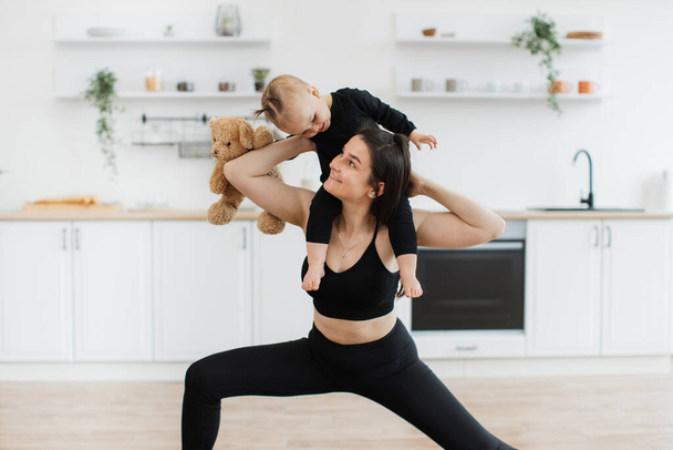 Attractive slim female keeping visual contact with kid held on shoulders while stretching in yoga position at home. Healthy mommy getting additional weights to body by carrying daughter and soft bear. - Фото, зображення