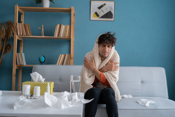 Ongelukkige jongeman gewikkeld in een deken met symptomen van verkoudheid of griep. Droevige zieke man heeft keelpijn en koorts thuis op de bank. Kopieerruimte - Foto, afbeelding
