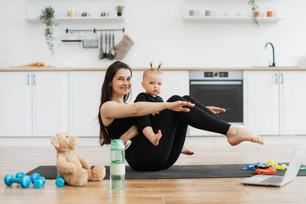 Cheerful young lady in sportswear holding boat pose with cute baby girl on laps in modern apartment. Sporty adult mom doing baby yoga with little daughter using online video on laptop at home. - Foto, immagini