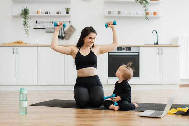 Athletic brunette woman and little girl lifting dumbbells while looking at each other on kitchen floor. Caring mother in activewear teaching inquisitive daughter strength exercises in apartment. - Fotografie, Obrázek