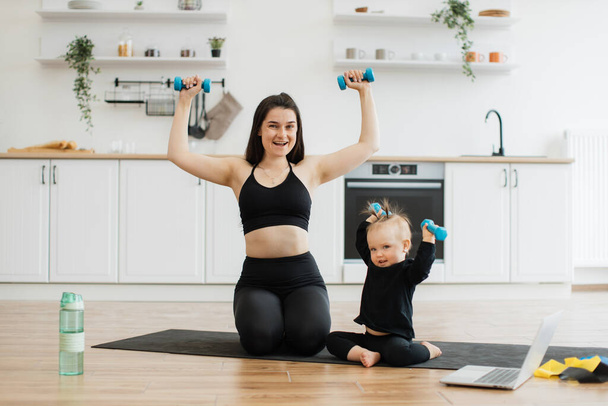 Emotional caucasian woman and kid in sports clothes raising weights with excitement while sitting on room floor. Happy young mom and baby girl exercising together with joy near computer at home. - Foto, imagen
