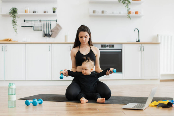 Attractive slim woman supporting little baby with dumbbells in hands while sitting on knees behind her on yoga mat. Loving mommy gently stretching daughters arms during fitness class at home. - Photo, Image