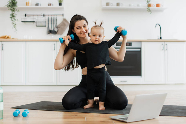 Full length view of little girl standing with weights in hands on yoga mat with smiling mother behind. Cheerful family woman combining childs entertainment with physical training in home interior. - Foto, immagini