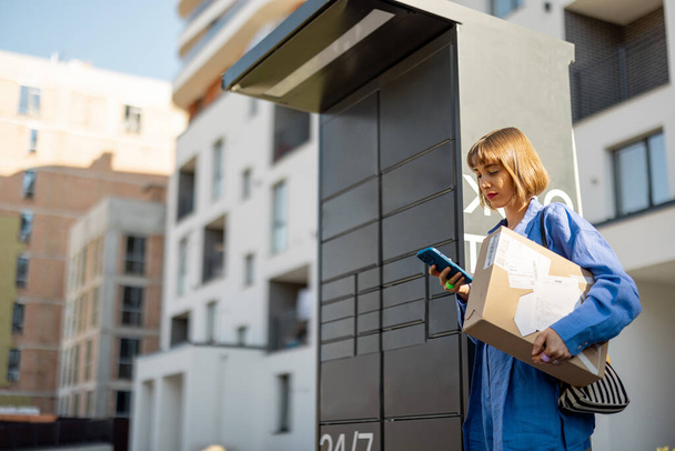 Mujer joven usando teléfono inteligente mientras está de pie con un paquete entregado con máquina de oficina de correos con taquillas automáticas. Nuevas tecnologías en el servicio de entrega, autoselección - Foto, imagen