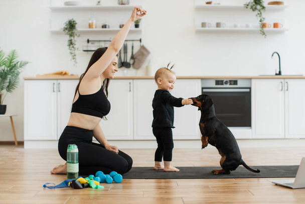 Active young woman and small kid in black yoga clothing teaching commands to smart animal while staying indoors. Delighted mother and daughter rewarding pets good behavior with homemade treats. - Photo, Image