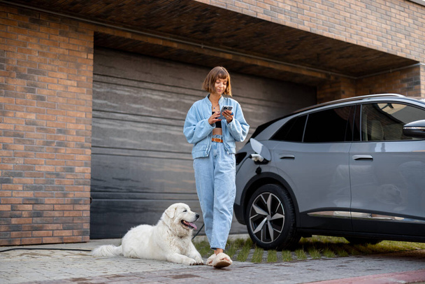 Молода жінка стоїть зі своїм собакою і використовує смартфон, заряджаючи електромобіль біля гаража свого будинку. Концепція сучасного способу життя та сталого розвитку - Фото, зображення