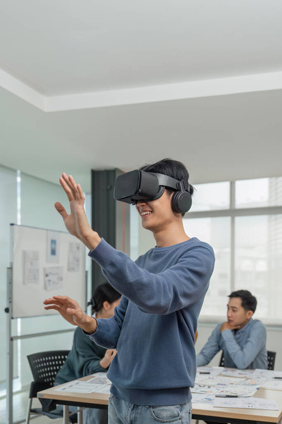 Un joven desarrollador asiático alegre y emocionado probó un nuevo juego de realidad virtual en un auricular de realidad virtual en la reunión con el equipo. Realidad virtual. - Foto, Imagen