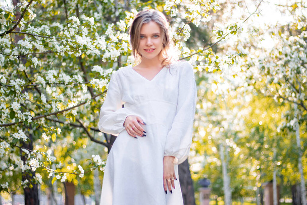 Portrait of a young woman in a white dress against the backdrop of blooming apple trees. The girl poses against the background of flowers in the spring park. - Foto, Bild