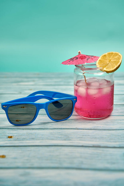 Refreshing summer drink with lots of ice an umbrella and a slice of lemon and blue sunglasses on a wooden table and a greenish background. - Foto, Imagen