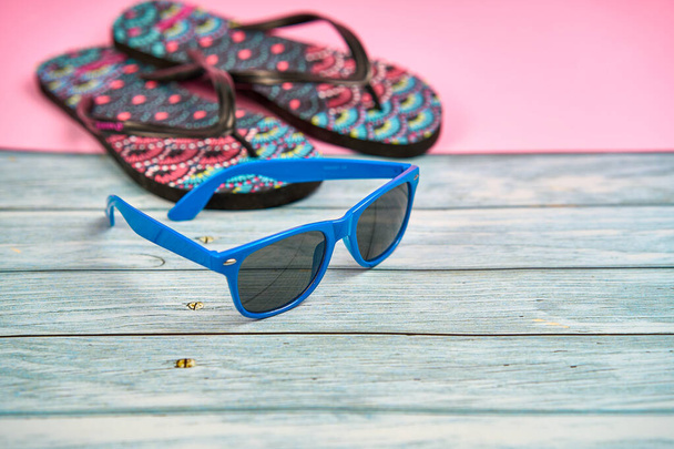 Blauwe zonnebril en teenslippers met een tropisch patroon op een houten tafel en een roze achtergrond. - Foto, afbeelding