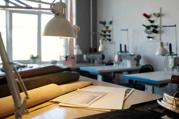 Zaměření na pracoviště koženého pracovníka s lampou, náčrtky a válcovanou koženou textilií různých barev proti psacím stolům s šicími stroji - Fotografie, Obrázek