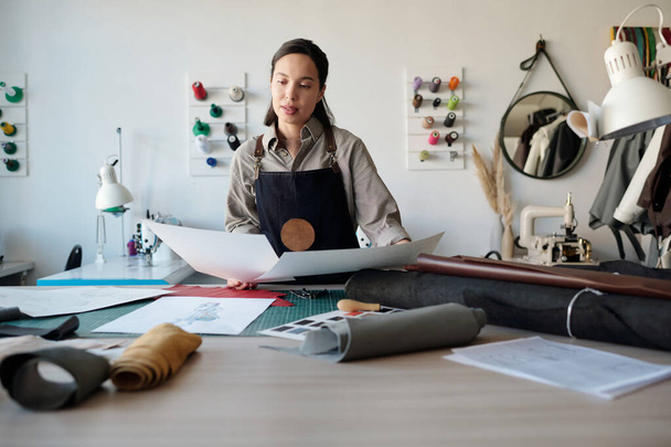 Молодая женщина в рабочей одежде, глядя на бумаги с эскизами в мастерской, выбирая новые модели одежды за столом с катаной кожей - Фото, изображение