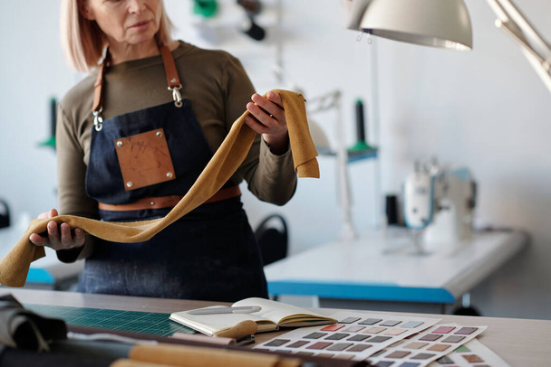 Gros plan du tailleur professionnel mature avec un morceau de cuir, daim ou autre textile de couleur jaune dans les mains debout par le lieu de travail en studio - Photo, image