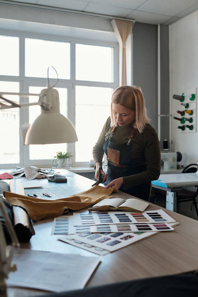 Blonde reife Schneiderin oder Modedesignerin schneidet Stück Leder am Arbeitsplatz mit Textilmustern, Skizzen und anderem Zubehör - Foto, Bild