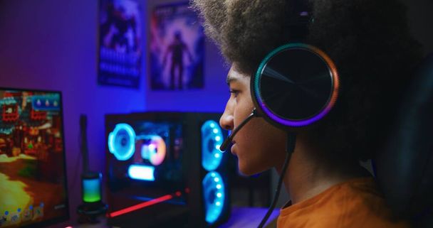 Happy gamer in headphones wins a round in multiplayer game on home personal computer. African American teenager plays third person shooter. Online video game live streaming or cybersport competition. - Foto, immagini