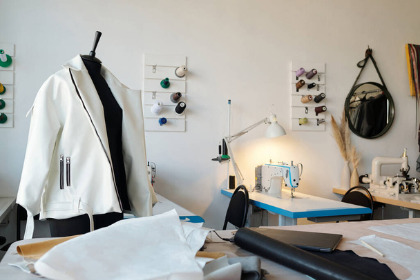 New white leather jacket on mannequin and workplace of tailor or leatherworker with spools of threads on wall and sewing equipment - 写真・画像