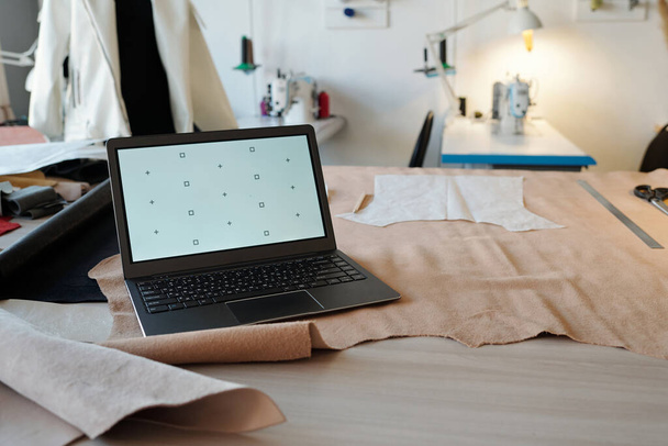 Werkplaats van looier of taillor met laptop op beige suède of leder textiel met papieren patronen tegen naaimachines - Foto, afbeelding