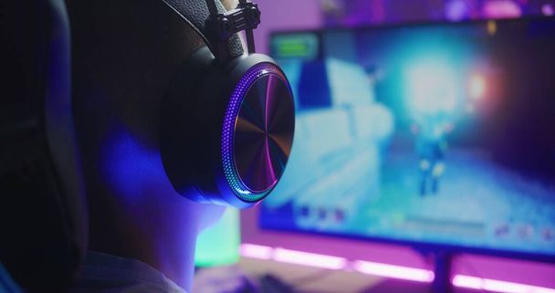 Young boy in headphones plays third person shooter. Gamer enjoys online video game on home personal computer. Desk illuminated by RGB LED strip light. Concept of gaming at home. Back view. Close up. - Photo, Image