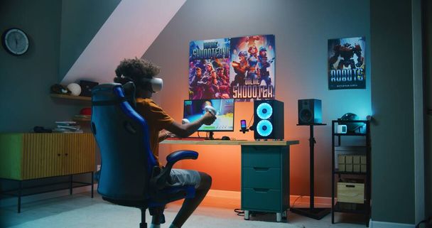 Un adolescent afro-américain dans un casque VR joue au jeu de tir virtuel en 3D à la troisième personne sur un ordinateur personnel à l'aide de contrôleurs sans fil. Jeu vidéo en streaming en ligne ou championnat de cybersport. Jeu à la maison. - Photo, image