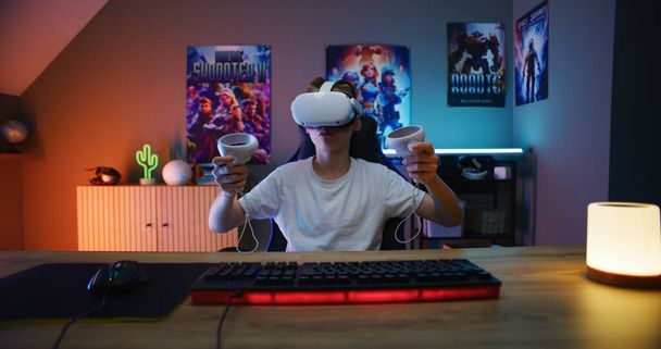 Fiatal játékos VR headset játszik virtuális online videojáték segítségével vezeték nélküli vezérlők. Szereti az e-sport bajnokságot. Meleg neon LED világítással ellátott szoba. Hazajárás. POV PC képernyő szemszögéből. - Fotó, kép