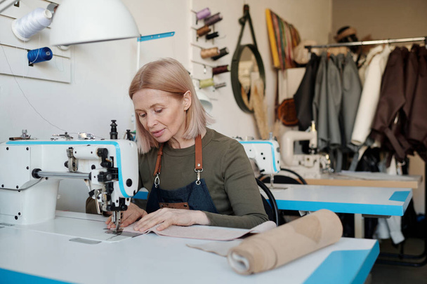 新しい革の服や流行のアイテムを縫製しながら、彼女のクライアントのための季節のファッションコレクションを作成する作業服の成熟した職人女性 - 写真・画像