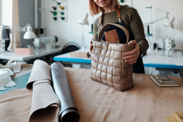 Cropped shot of experienced leatherworker holding new stylish leather handbag while standing by workplace with beige suede textile - Fotografie, Obrázek