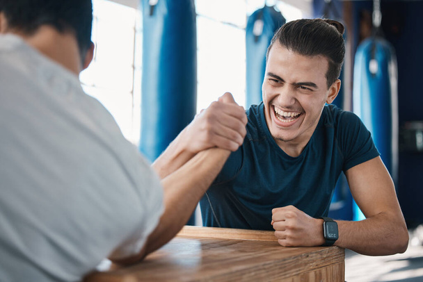 Δυνατός, δραστήριος και οι άνδρες χέρι πάλη στο γυμναστήριο σε ένα τραπέζι, ενώ είναι παιχνιδιάρικο για μια πρόκληση. Ανταγωνιστικότητα, παιχνίδι και άνδρες ή αθλητές που δίνουν μάχη μυών δύναμης για διασκέδαση, δέσιμο και φιλία - Φωτογραφία, εικόνα