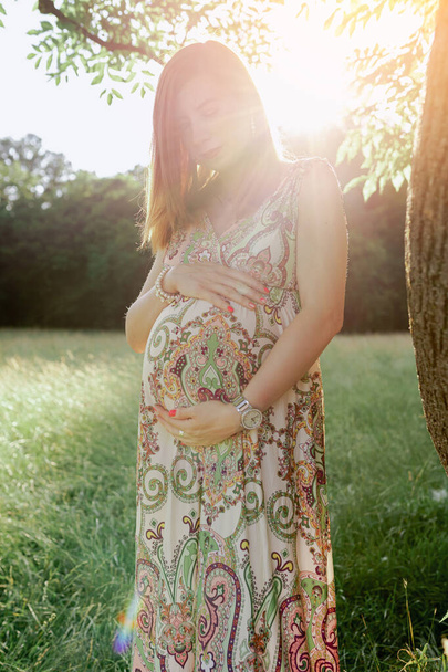 Portrait attractive red-haired young woman, pregnant, in summer dress, holding belly with hand, against blurred field background - Fotografie, Obrázek