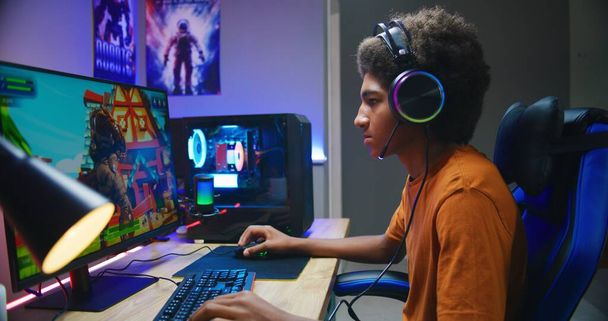 Professional eSports African American young gamer puts on headphones and plays online third-person 3D shooter on modern PC in stylish neon room. Video game live streaming or cybersport competition. - Photo, image