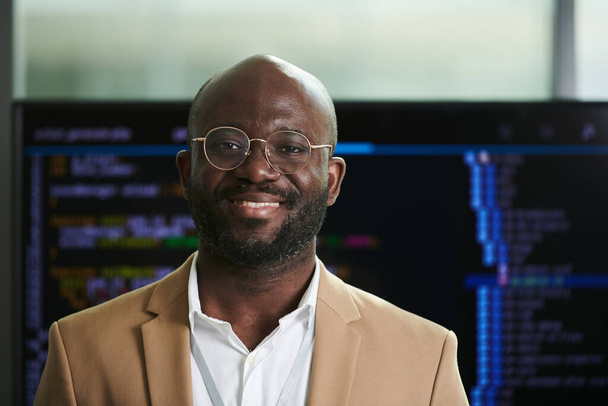 Young smiling African American businessman in eyeglasses and formalwear looking at camera while standing against interactive board with data - Foto, afbeelding
