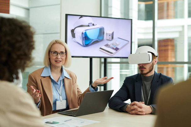 Confident mature business coach explaining something to employees at working meeting while making presentation of new vr headset - Фото, изображение